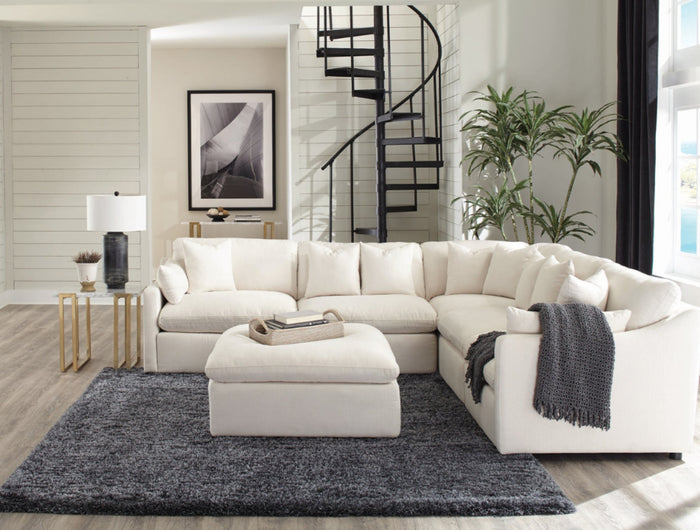 A cozy living room with a light gray sectional sofa, wooden shelving units filled with books, framed photos on walls, and soft lighting from lamps. Carpeted floor and stairs leading up in the background.