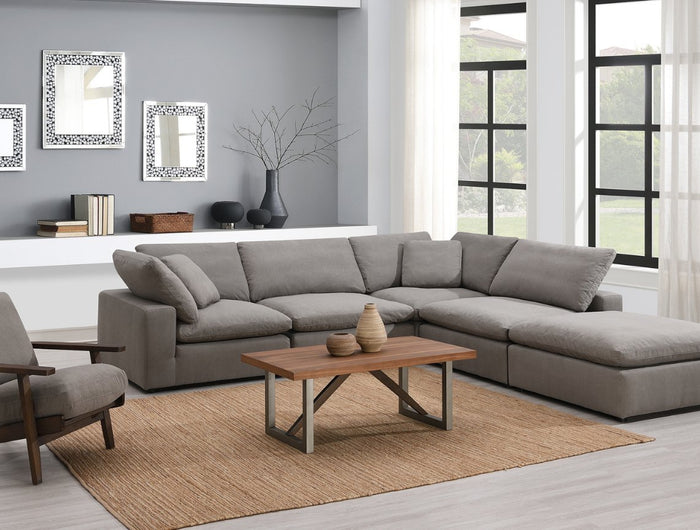 Modern living room with a gray sectional sofa, wooden armchair, and a wooden coffee table on a beige rug. Large windows with gauzy curtains let in natural light. Three frames and a vase on a white shelf accent the wall.