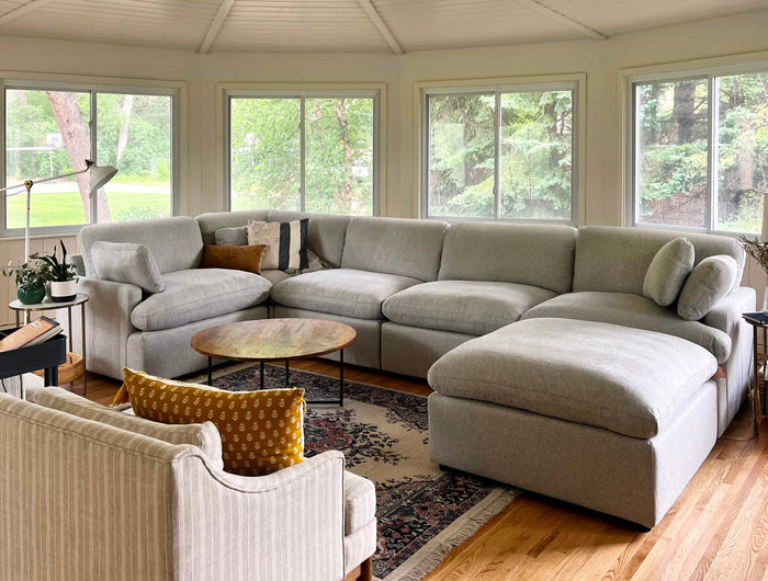 A cozy living room with a large gray sectional sofa, a round wooden coffee table, a patterned area rug, and an armchair with a mustard cushion. The space is bright with large windows showing greenery outside.