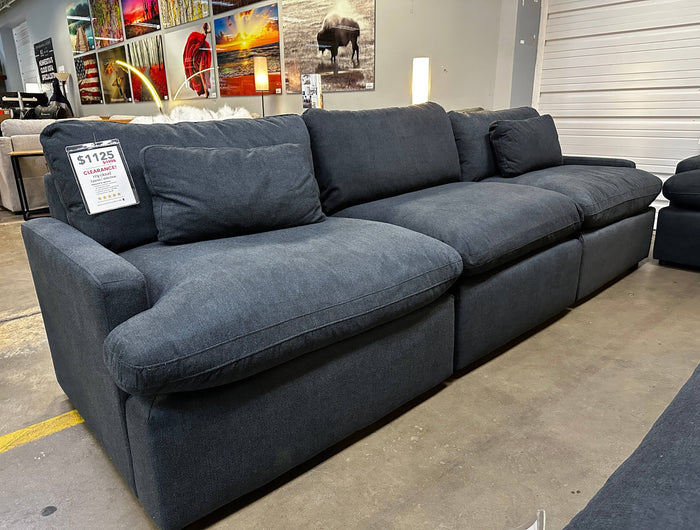 A large, dark gray sectional sofa with plush cushions is on display in a showroom. A price tag shows $1125 for clearance. Various framed art pieces are visible on the wall behind the sofa.