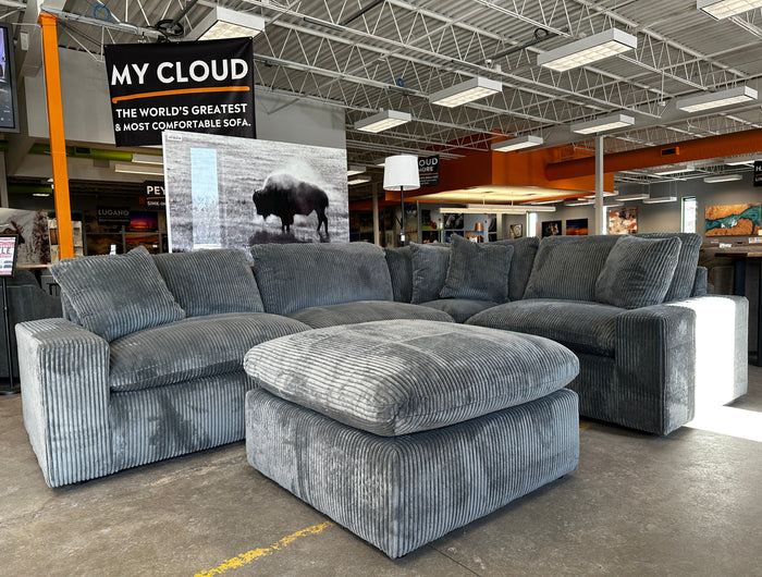 A large, plush gray sectional sofa with a matching ottoman is displayed in a showroom. A sign above reads, MY CLOUD: The Worlds Greatest & Most Comfortable Sofa. A bison image is in the background.