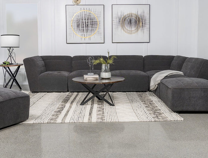 A modern living room with a dark gray sectional sofa, a round coffee table, and a patterned rug. Two abstract paintings and a geometric chandelier adorn the wall. Plants and a floor lamp complete the decor.