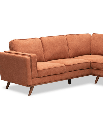 The Maddox LSF is a rust-colored L-shaped sofa with wooden legs and plush cushions. It boasts a modern, mid-century design with uniform texture and sleek looks, set against a plain white background.