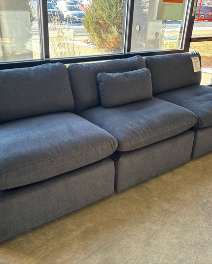 A large 3-piece My Cloud Slate Blue sectional sofa with plush cushions and a small rectangular throw pillow sits in a room with concrete flooring. Natural light enhances the sofas texture and highlights its machine washable design.