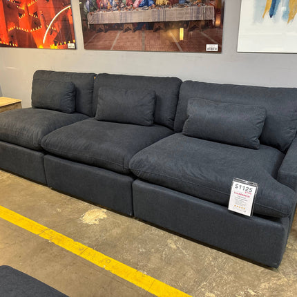 The 3 Piece My Cloud Slate Blue sectional sofa, featuring subtle stitching and plush cushions, is priced at $1125. It sits against a gray wall adorned with colorful artwork, beside a wooden side table with ornate legs on the concrete floor.