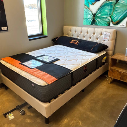 In a showroom, a Full Size Tufted Cream Headboard & Frame (floor sample) holds a full-size mattress topped with a large black pillow featuring orange RCL letters. A wooden nightstand complements the setup, while sunlight and artwork enhance the inviting display of our product line.