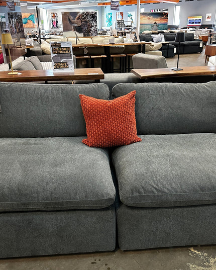 The 2 Piece My Cloud Armless Classic Gray sofa with a red textured pillow is displayed in a furniture store, surrounded by new pieces like sofas, tables, and decorations. A price tag is visible on the armrest in the spacious showroom.