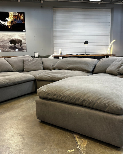 The 4 Piece + Ottoman Peyton Gray, a spacious sectional sofa in excellent condition with plush cushions, is showcased on the polished concrete floor of a showroom. Colorful artworks adorn the wall behind, and some furnishings display visible price tags.