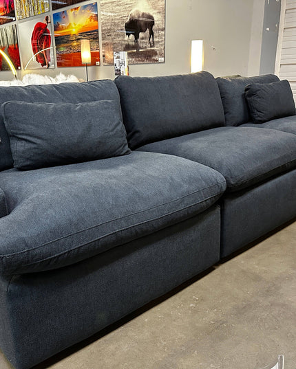 A 3 Piece My Cloud Slate Blue sectional sofa with dark gray upholstery and plush cushions is displayed in a furniture store. A sale tag shows a reduced price of $1125. Bright, colorful artwork hangs on the wall in the background, highlighting the sofas elegant design.