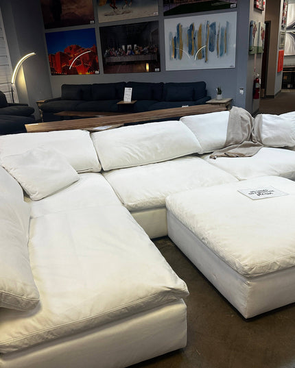 A 4 Piece + Ottoman Peyton White sectional with a right arm, in good condition as a floor model, sits in a furniture store. It’s surrounded by other furniture pieces and wall art, with polished concrete flooring and warm ambient lighting enhancing the display.