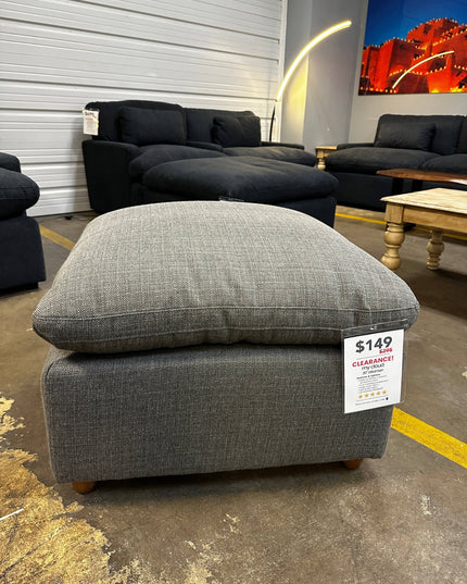 A My Cloud Slope 30 Stone Ottoman, a showroom floor model with discontinued fabric and priced at $149 clearance, sits in the display. Only one is available, surrounded by a black couch, wooden table, modern floor lamp, and a large photograph on the wall.