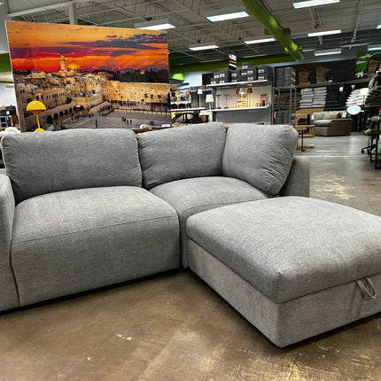 The showroom features a 3 Piece Lugano Gray Sectional with a chaise and ottoman for $595. In the background, large cityscape wall art depicts a sunset, surrounded by various other furniture pieces.