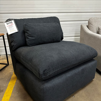 The My Cloud Slate Blue Armless Chair, priced at $295, is on display on a warehouse floor. Despite its damaged packaging, the chair appears like new. Nearby are a light-colored couch and a small table, partially visible.