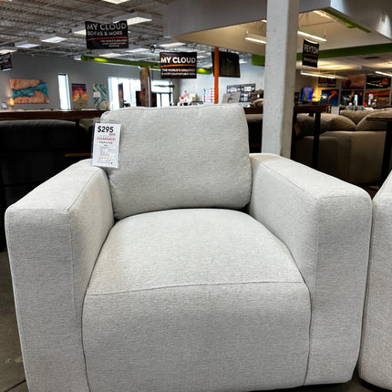 The Lugano Linen Chair, an open box item with damaged packaging, sits comfortably in a furniture store priced at $295. Its wide arms and like-new appearance stand out amidst other sofas and signage under the high ceilings and lighting.