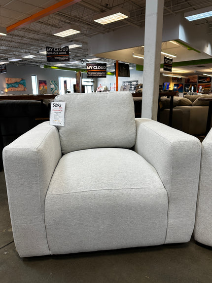 The Lugano Linen Chair, an open box item with damaged packaging, sits comfortably in a furniture store priced at $295. Its wide arms and like-new appearance stand out amidst other sofas and signage under the high ceilings and lighting.