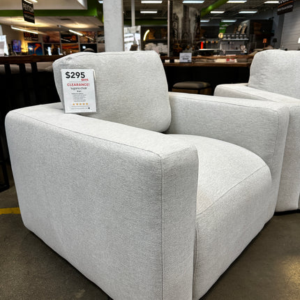 The Lugano Linen Chair, an open box comfy light gray armchair with a modern boxy design, is showcased in a store. Despite its clearance price of $295 due to damaged packaging, it appears new against a backdrop of other furniture and the stores ambiance.