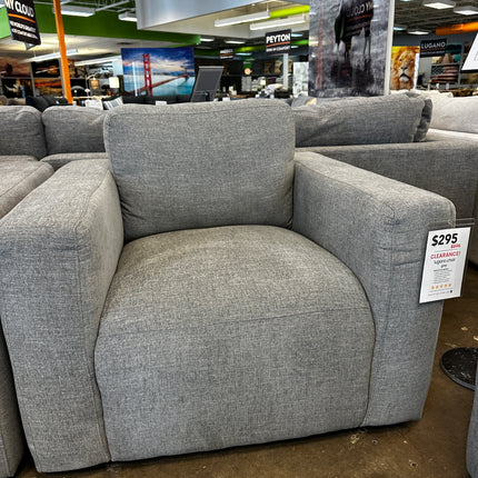 The Lugano Gray Chair, in open-box condition and priced at $295, showcases wide armrests and a cushioned backrest. Its displayed in a stores clearance section on a polished concrete floor among other furniture.