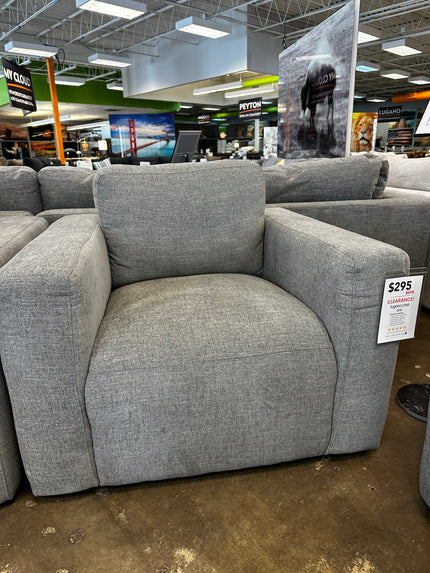 The Lugano Gray Chair, in open-box condition and priced at $295, showcases wide armrests and a cushioned backrest. Its displayed in a stores clearance section on a polished concrete floor among other furniture.