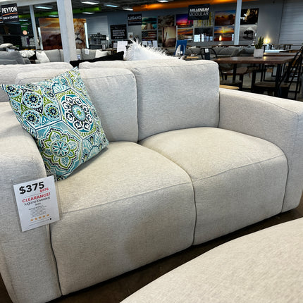 The Lugano Linen Loveseat, a beige floor model, accompanied by a colorful geometric pillow, is showcased in a furniture store. Priced at $375 on clearance, its surrounded by rows of tables and chairs under bright lighting.