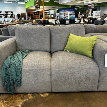 The Lugano Gray Loveseat is displayed in a showroom with a green throw pillow and teal blanket. Despite its inviting appearance, its priced at $375 due to damaged packaging.