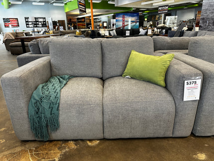 The Lugano Gray Loveseat is displayed in a showroom with a green throw pillow and teal blanket. Despite its inviting appearance, its priced at $375 due to damaged packaging.