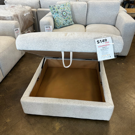 The Lugano Linen Storage Ottoman, a beige fabric piece with an open lid offering cozy storage, sits on the store floor beside a light grey sofa with a decorative pillow. Its marked with a clearance price of $149.