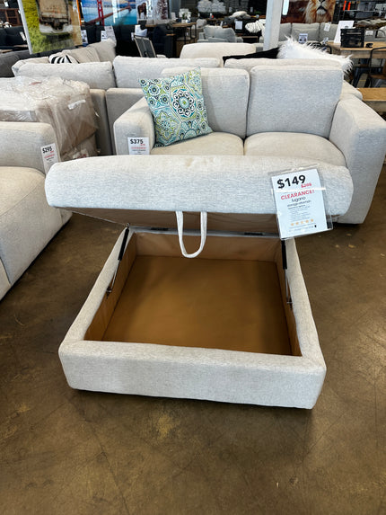 The Lugano Linen Storage Ottoman, a beige fabric piece with an open lid offering cozy storage, sits on the store floor beside a light grey sofa with a decorative pillow. Its marked with a clearance price of $149.