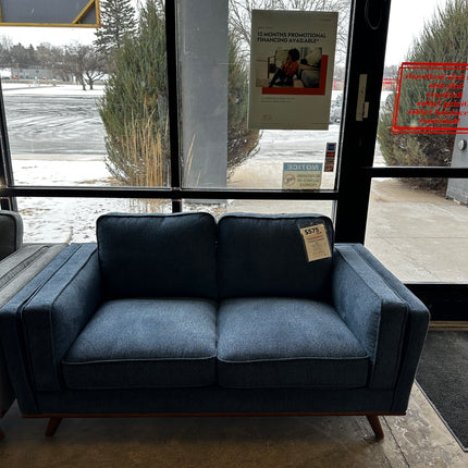 The Maddox Blue Loveseat, a two-seater with wooden legs, is showcased near a glass door. Though its packaging shows slight wear, a promotional poster adds charm against the snowy backdrop as the price tag hangs from its cushion.