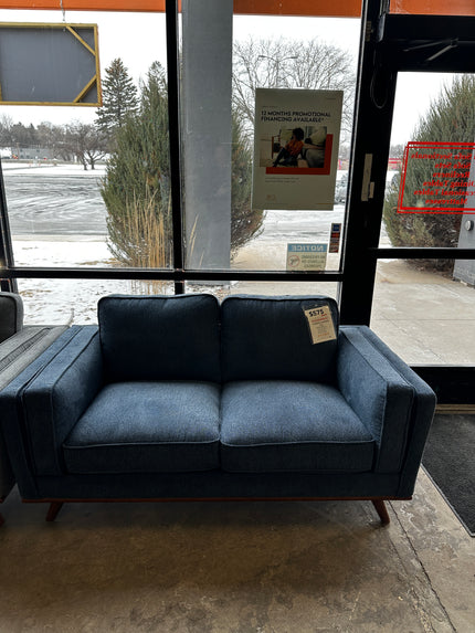 The Maddox Blue Loveseat, a two-seater with wooden legs, is showcased near a glass door. Though its packaging shows slight wear, a promotional poster adds charm against the snowy backdrop as the price tag hangs from its cushion.