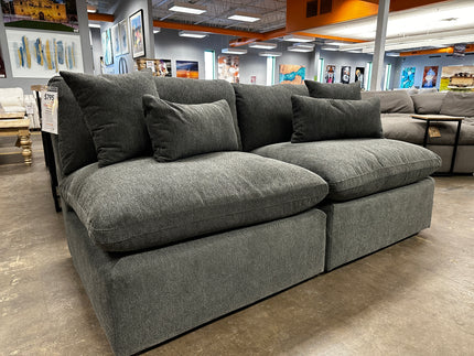 The 2 Piece My Cloud Armless Loveseat in Classic Gray, featuring deep, machine washable cushions and matching pillows, is displayed in a furniture store. It sits against vibrant paintings on a polished concrete floor under sleek lighting.