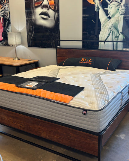 A showroom display features the My Cloud Mattress 400 Luxe King / Floor Model with a wooden bed frame, adorned with black and orange pillows and a folded black mat. Behind it are posters of a tiger and the Statue of Liberty, complemented by a nearby wooden desk and chair.