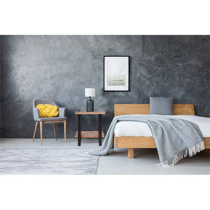 A minimalist bedroom exudes rustic charm with a wooden bed, gray bedding, and a light gray throw. A Live Edge End Table with a lamp sits beside the bed. Nearby, a chair is accented by a yellow cushion. Textured gray walls are enhanced by an elegant framed picture above.