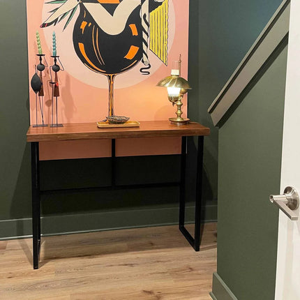 The Rosewood Console with black metal legs stands beneath a colorful abstract bird and plant wall art. Surrounded by dining room furniture, it holds a decorative sculpture, tray, and metallic lamp. A staircase railing is visible on the right.